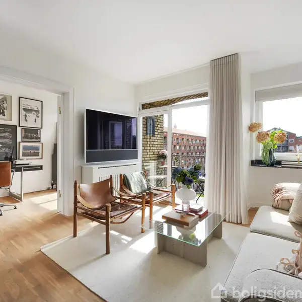Stue med sofa og lænestole, vendt mod et sofabord af glas. Et fjernsyn hænger på væggen. Sollys strømmer ind fra vinduerne, og en kontorplads ses i baggrunden.