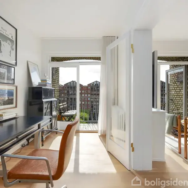 Et moderne kontor med et sort skrivebord og brun stol, dekoreret med kunstværker på væggen, forbindes til opholdsstue med glasdøre, med udsigt til en balkon og bygninger udenfor.