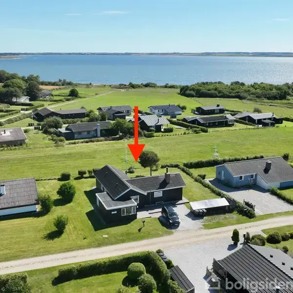 Et hus med grå tagsten ligger i en række af enfamiliehuse. En rød pil peger på huset. I baggrunden ses en sø eller hav, omgivet af marker.
