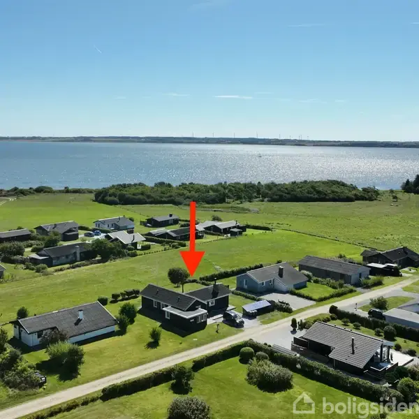 Hus med sort tag står i landligt område tæt på vandet. En rød pil peger på huset, omgivet af grønne marker og andre huse i nærheden, ved kysten.