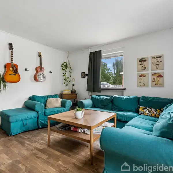 Turkis sofa i hjørne, med to guitarer på væggen. Stuebord i træ i midten. Grønne planter og vindue med udsigt til have. Fire dekorationer på væggen.