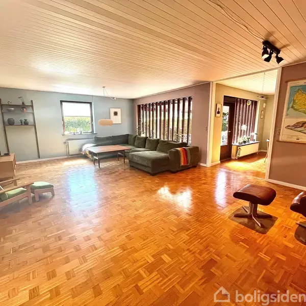 En grøn sofa står i en stue med et træbord foran. En hvid lampe hænger over bordet. Store vinduer viser udsigt til en frodig terrasse.