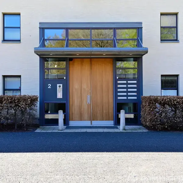 En moderne bygning med en stor indgangsdør omgivet af glaspartier og små vinduer. Tallet "2" er synligt ved siden af døren. Buske flankerer indgangen, beliggende foran en asfalteret vej.