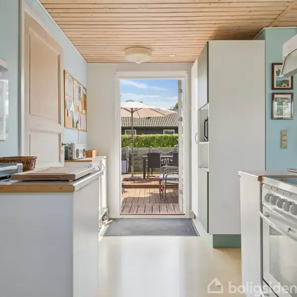 Et køkken med hvide skabe og apparater åbner op ud til en terrasse. Der er en bordplade, en stegeplade og rektangel loftslampe. Udenfor ses et bord og stole under en parasol.