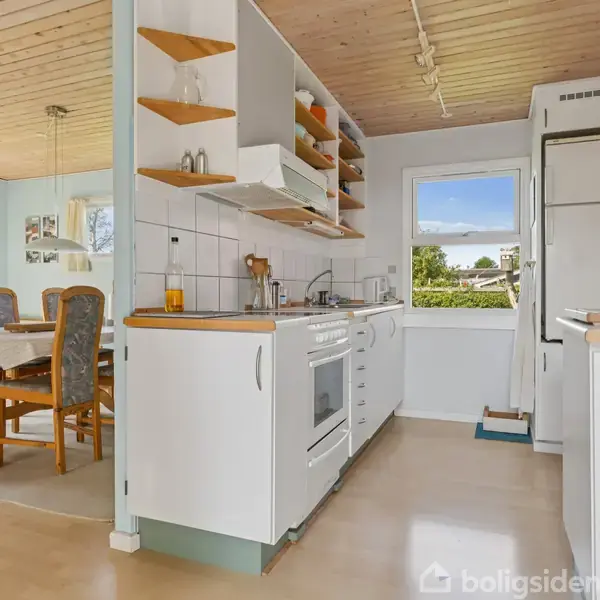 Køkkenet er funktionelt med hvidt interiør, åben hyldeopbevaring og komfur. Det ligger i et lyst rum, der åbner op til en spisestue med spisebord og fem stole.