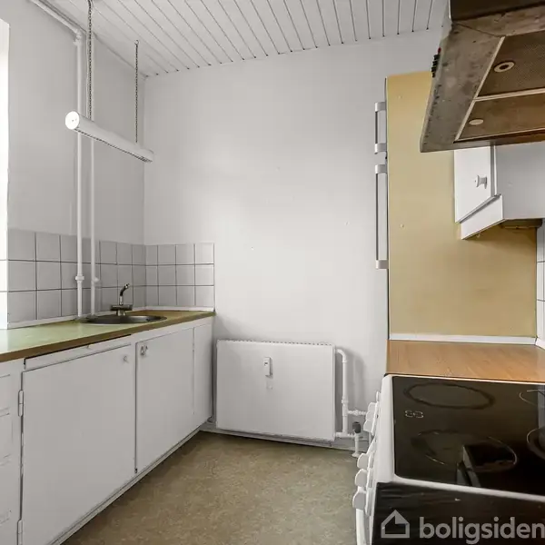 Et lille køkken med hvide skabe og skuffumskuffer. Komfur står ved højre væg, vask ved venstre vindue. Fliser dekorerer væggen bag vask og komfur. Ingen tekst er fundet.