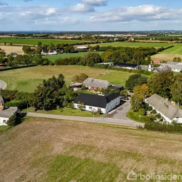 Flere huse ligger spredt i et åbent, landligt landskab med marker, træer og veje. Bebyggelsen er omgivet af grønne områder og landbrugsjord under en delvist skyet himmel.