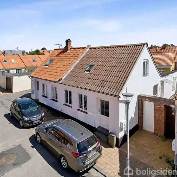 Et hvidt hus med rødt tegltag står ved en vej, hvor to biler er parkeret foran. Omgivet af lignende tage i et villakvarter. Et blåt parkeringsskilt ses tæt på huset.