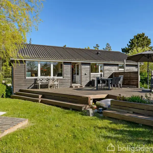 Et træhus står på en grøn græsplæne. På terrassen er der et bord med stole samt en parasol. Haven er omringet af træer og buske i et solrigt område.