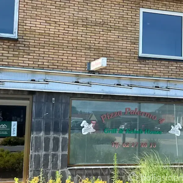 Et pizzeria med navnet "Pizza Palermo 2" vises. Skiltet på vinduet siger "Grill & Kebab House" og inkluderer telefonnummeret "Tlf: 92 27 20 16". Bygningens facade er af mursten.