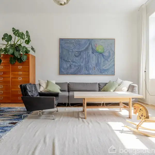 Sofaen står i en lys stue med en træbord foran. En sort lænestol og en gyngestol er ved siden af. Store grønne planter står på en kommode.