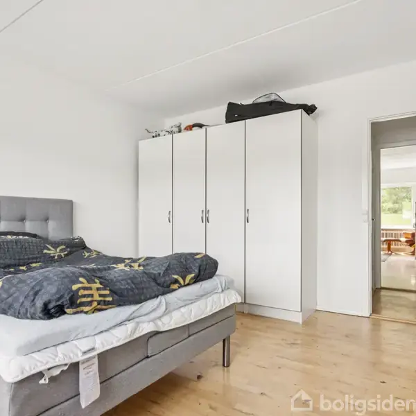 En seng med gråt sengegærde står i et soveværelse ved siden af en hvid garderobe. Rummet har trægulv og en åben dør viser adgang til en opholdsstue.