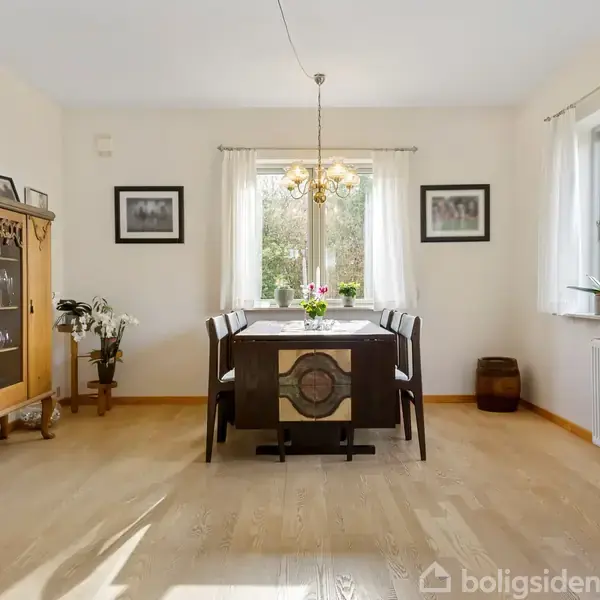 Et spisebord er centralt placeret i et lyst rum. Det er omgivet af stole og dekoreret med blomster og en lysekrone. Store vinduer og træmøbler præger omgivelserne.