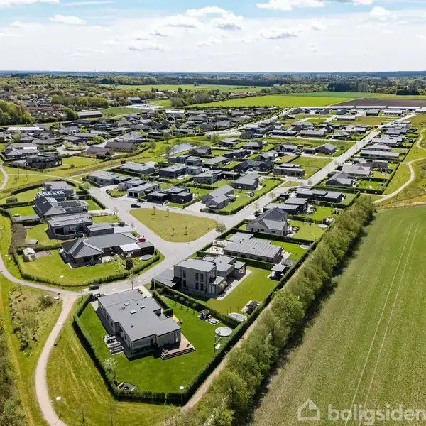 En lille sø omgivet af græsarealer ligger centralt i et nybygget boligområde med huse i rækker. Rundkørslen markerer indgangen til kvarteret, der omringes af åbne marker.