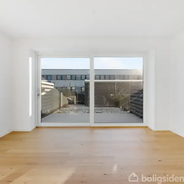 Et tomt rum med trægulv har store glasdøre. Sollys skinner ind, og udenfor ses en terrasse med et skur og et nabohus i baggrunden.