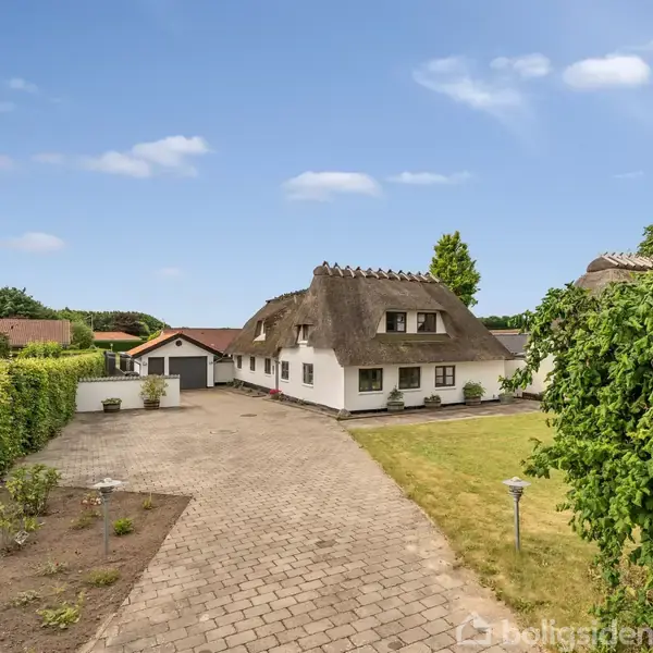 Hus med stråtag står stille på brostensbelagt indkørsel omgivet af velholdt have og grønne hække under en blå himmel med lette skyer.