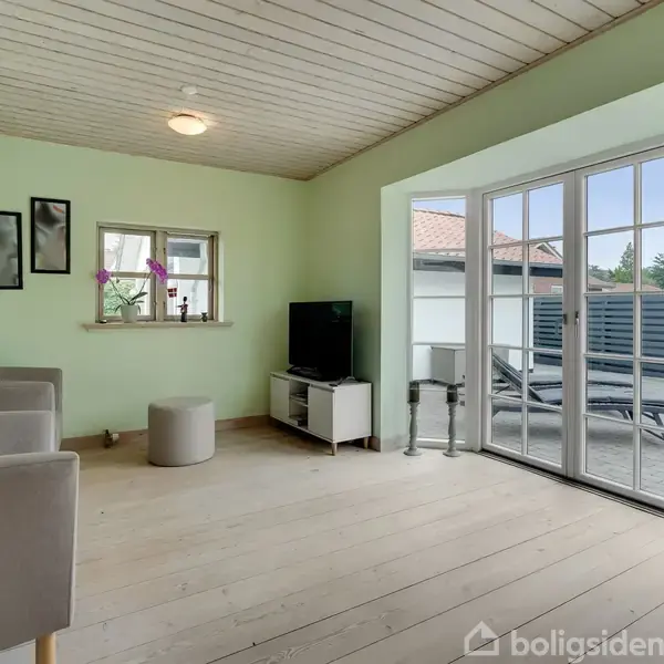 Stue med lysegrønne vægge indeholder to grå lænestole, et lille bord, og et TV. Store glasdøre fører ud til en terrasse med liggestole. To billeder hænger på væggen.