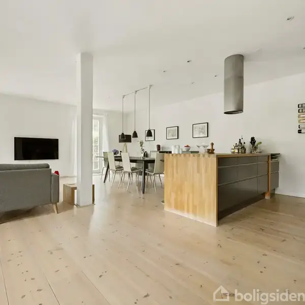 Stue med åbent køkken, træbord og stole. En grå sofa står ved væg med fladskærms-tv. Store vinduer giver lysindfald, moderne køkkenø centralt placeret. Ingen tekst i billedet.