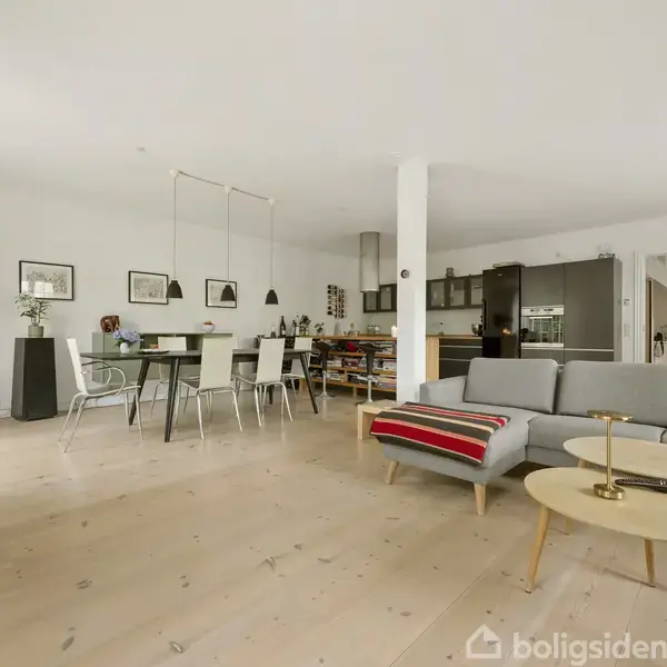 Stue med gråt hjørnesofa som står på lyst trægulv, to runde borde foran. Spisebord med stole i baggrunden. Væggene er hvide, og rummet er lyst og rummeligt.