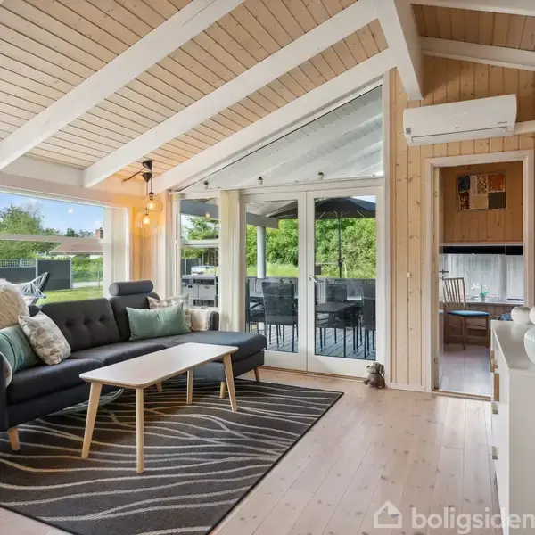En moderne stue med en mørkegrå sofa og træbord. Store vinduer slipper lys ind. Udenfor ses en terrasse med havemøbler. Loftet har synlige bjælker, og der hænger en lampe.