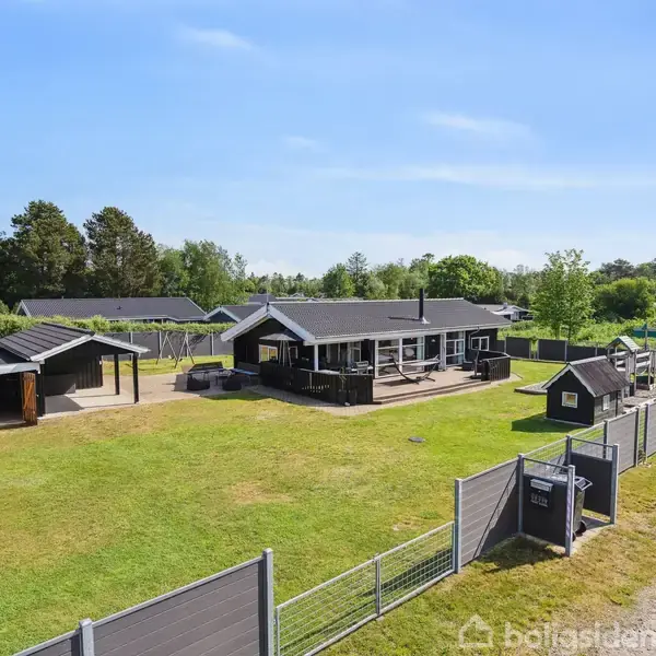 En sortmalet sommerhus med terrasse ligger i en indhegnet have med græs og træer. Til venstre ses en garage, og der er et skilt i forgrunden.