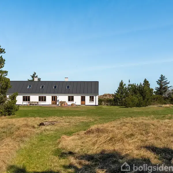 Et hvidt hus med sort tag står midt på en grøn græsplæne omgivet af buske og træer under en blå himmel.