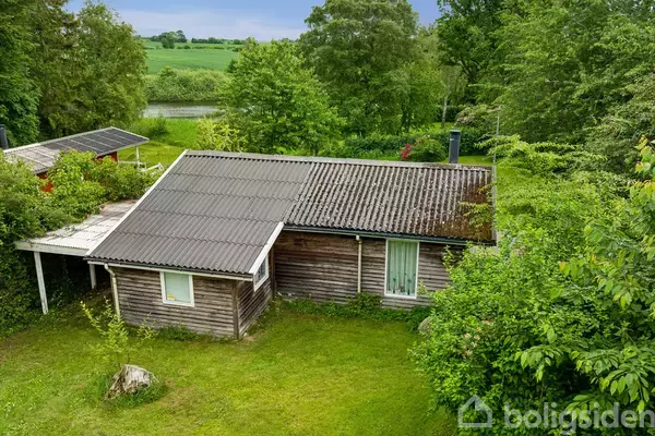 Et lille træhus står stille omgivet af frodig grøn beplantning. Taget har både forskudte og tilgroede områder. I baggrunden ses en åben mark med træer.