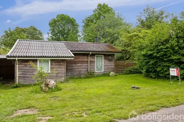 Et lille træhus står stille omgivet af grønne buske og græsplæne. Foran huset er et skilt med teksten "Til salg".
