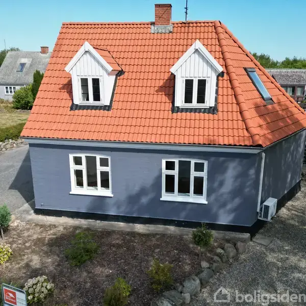 En lille træbro står ved en dam omgivet af højt græs og træer i en frodig naturpark. Grussti løber foran scenen.