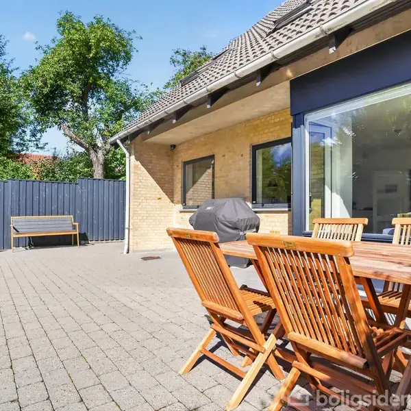Træbord og stole står på en flisebelagt terrasse ved siden af et hus med gule mursten. I baggrunden ses en bænk, et plankeværk, og grønne træer.