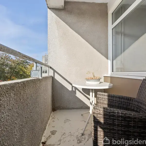 En balkon med et rundt bord og en fletstol. Bordet har en lille potteplante. Sollys skinner på betongulvet, og der er udsigt til træer og blå himmel.
