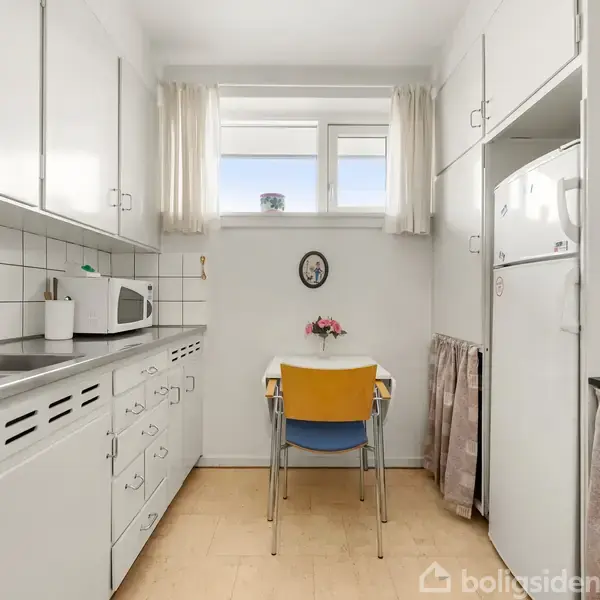 Et lyst køkken med hvide skabe og en lang bordplade. Et lille bord med to stole står ved et vindue med gardiner, og en mikroovn er placeret på bordpladen.