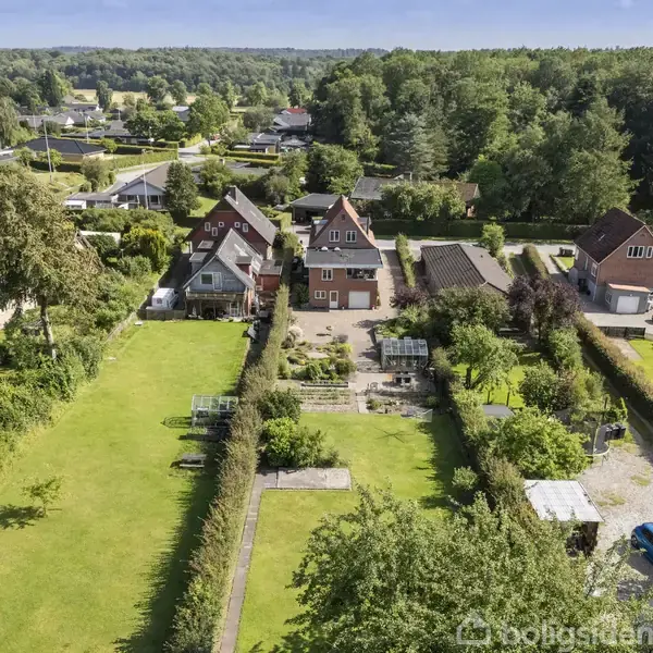 Huse med stejle tage står langs en vej omgivet af frodige haver og grønne plæner. En blå bil ses parkeret til højre. Skovområde i baggrunden.