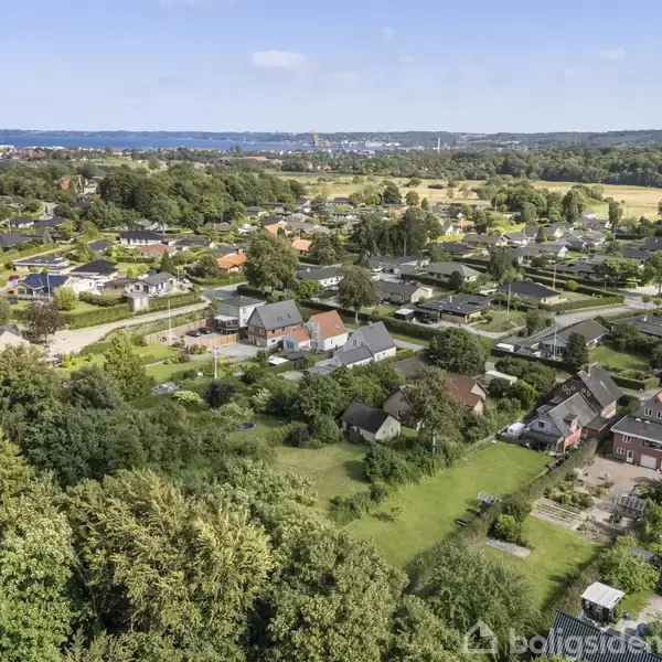 Boligområde med huse spredt ud over grønne landskaber, set fra oven. I baggrunden ses en kystlinje og hav. Landskabet er frodigt med træer og marker.