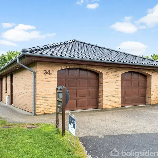 En gul murstensgarage med sort tegltag har to brune porte og nummer 34 på siden. Omgivet af grønne hække og græsplæne med en tilsluttet indkørsel. En skilt er foran garagen.