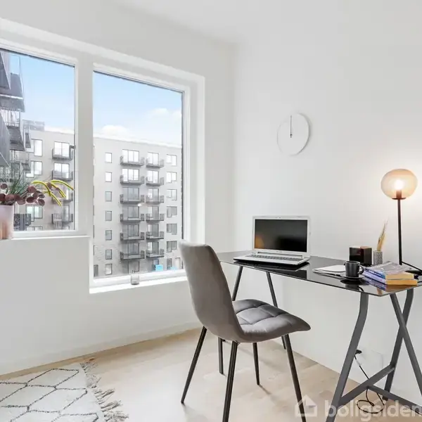 En minimalistisk skrivebordsscene med en bærbar computer tændt, omgivet af notesbøger og en lampe. Designstol foran, og vinduer viser lejlighedsbygninger udenfor. En plante er placeret ved vinduet.