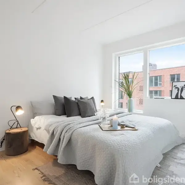 En dobbeltseng er redt med gråt sengetøj, omgivet af en natlampe og en plante ved vinduet i et lyst soveværelse. Møblerne er stilfulde og minimalistiske.