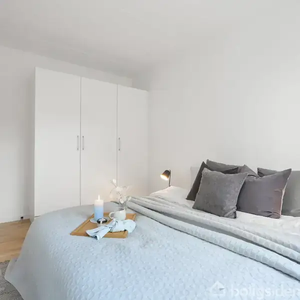 Soveværelse med en stor seng dækket af et lyseblåt tæppe og puder. En bakke med lys står på sengen. Hvidt klædeskab i baggrunden ved siden af en åben dør.