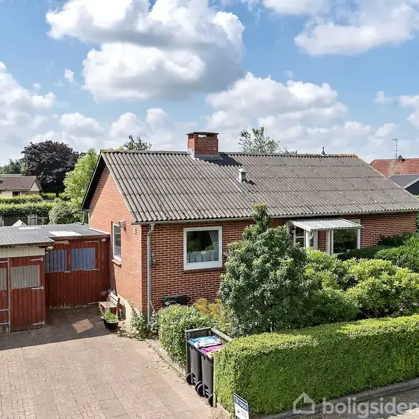 Rødt murstenshus med et gråt tag ligger i en velplejet have. Foran huset er der en flisebelagt indkørsel, og en garagebygning er til venstre. Skyfri himmel med nogle skyer.