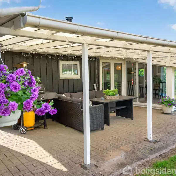 Overdækket terrasse med mørk sofa og bord. Blomster i potter hænger på væggen. Terrassegulvet er af mursten, og der vokser græs omkring. Vejret er solrigt med blå himmel.