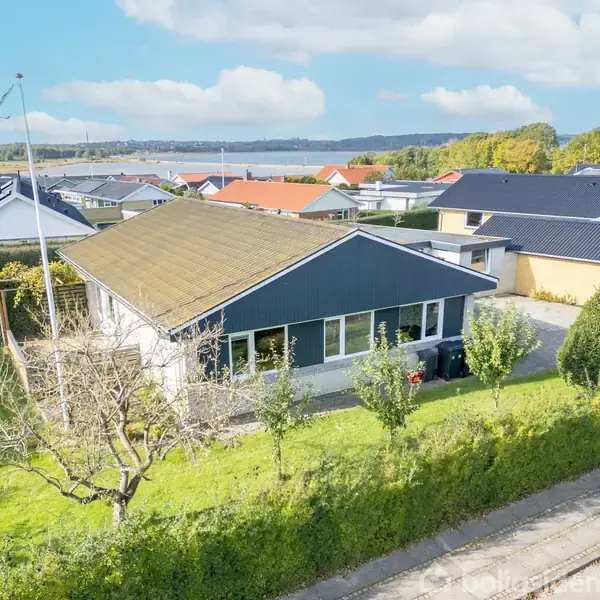 Et enkelt hus med gavltag står på en velplejet græsplæne omkranset af hække. Der er en flagstang ved siden af huset i et villakvarter med udsigt over vandet.
