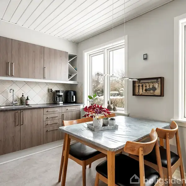 Et moderne køkken med trækøkkenskabe og en køkkenvask. En spisebord med fire træstole står ved et vindue med udsigt til træer. En plante og et maleri pynter rummet.