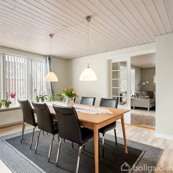 Spisebord med seks stole står i en lys spisestue med trægulv. Hængende lamper lyser over bordet. Store vinduer med gardiner og planter skaber en hyggelig atmosfære.