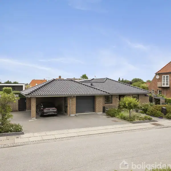 Murstensvilla med mørkt tag står ved vejen. En bil er parkeret under den åbne garage. Omkringliggende haver har grønne buske og blomsterbede. Klar blå himmel overhead.