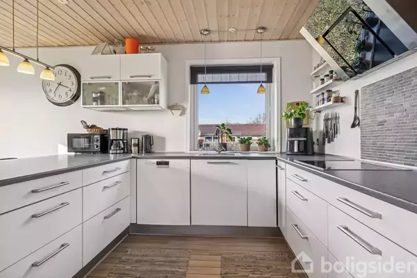 Et køkken med hvide skabe og moderne apparater. En kaffemaskine og mikroovn står på bordpladen. Et vindue med udsigt til et hus omgivet af planter. Klokke på væggen.