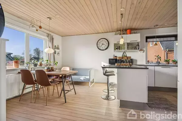 Et lyst køkken-alrum med træloft har et rundt spisebord med fire stole. Ved siden af er et moderne køkken med en væghængt hylde. Store vinduer giver udsigt til en have.