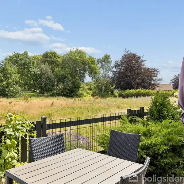 Terrassebord med stole står stille i en have omgivet af grønne planter og hegn. I baggrunden ses en åben mark med spredte træer under en blå himmel med skyer.