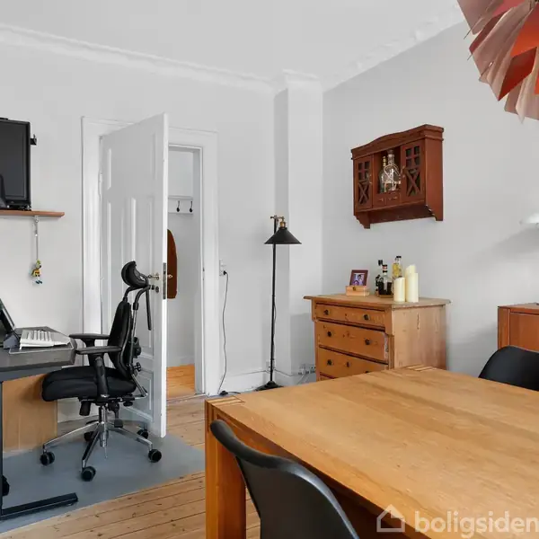 Et kontor med skrivebord udstyret med computer og kontorstol. Til højre en spisebord i træ. Rummet har lyse vægge og flere træmøbler, inklusive en kommode og en skænk.