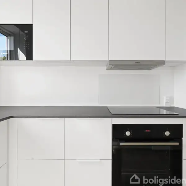Et moderne køkken med hvide skabe og sorte bordplader. Der er en indbygget sort ovn og en kogeplade. Et vindue ses i spejling ved mikrobølgeovnen.
