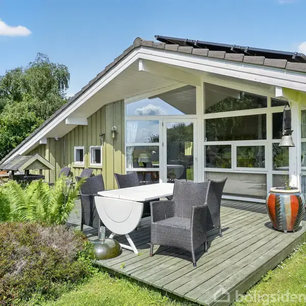 En velplejet have med græsplæne og buske omkranser et hus med store vinduer. I haven står en tørrestativ og et fuglebad. Skyfri himmel i baggrunden.